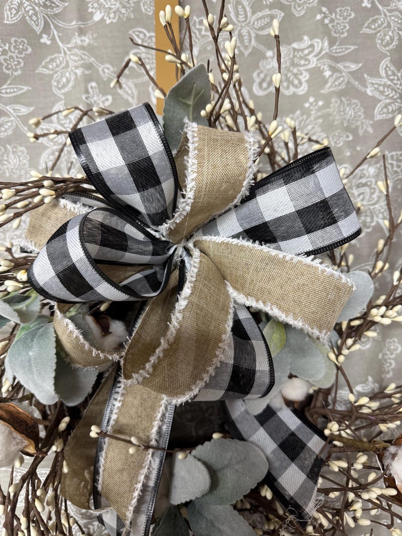 May include: A close-up of a decorative wreath featuring a large bow with black and white checkered ribbon and natural burlap ribbon. The wreath includes sprigs of white berries, green leaves, and cotton bolls, creating a rustic, farmhouse aesthetic.