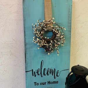May include: A teal blue wooden sign with a white and brown wreath hanging from a burlap ribbon. The sign says "welcome to our home" in black lettering.