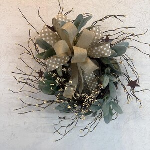 May include: A decorative wreath featuring a large tan bow with white polka dots and a smaller tan bow. The wreath includes green leaves, brown twigs with small white buds, and brown star accents. The wreath is designed for home decor.