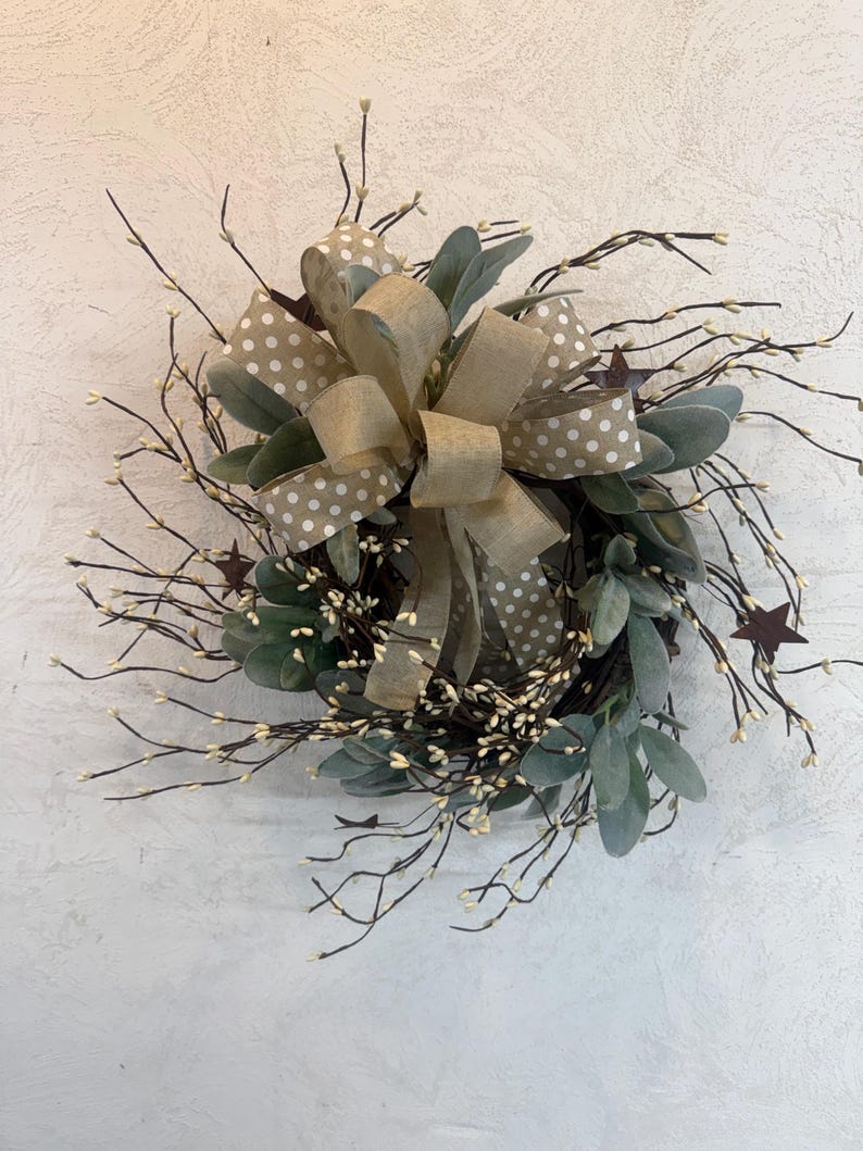 May include: A decorative wreath featuring a large tan bow with white polka dots and a smaller tan bow. The wreath includes green leaves, brown twigs with small white buds, and brown star accents. The wreath is designed for home decor.
