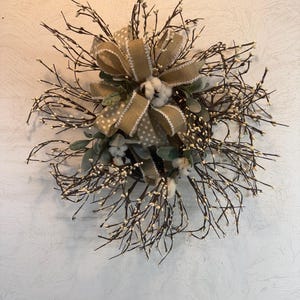 May include: A rustic wreath featuring a large burlap bow with a polka dot pattern, cotton accents, and sprigs of white berries. The wreath is made of dark brown twigs and green leaves, creating a natural, farmhouse aesthetic.