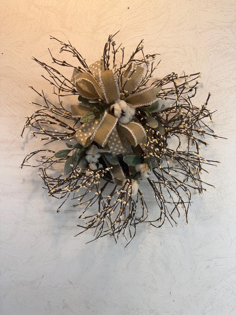 May include: A rustic wreath featuring a large burlap bow with a polka dot pattern, cotton accents, and sprigs of white berries. The wreath is made of dark brown twigs and green leaves, creating a natural, farmhouse aesthetic.