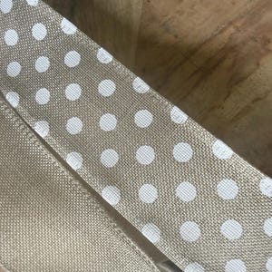 May include: Close-up of tan ribbon with white polka dots. The ribbon is made of a textured fabric and is folded over itself. The background is a wooden surface.