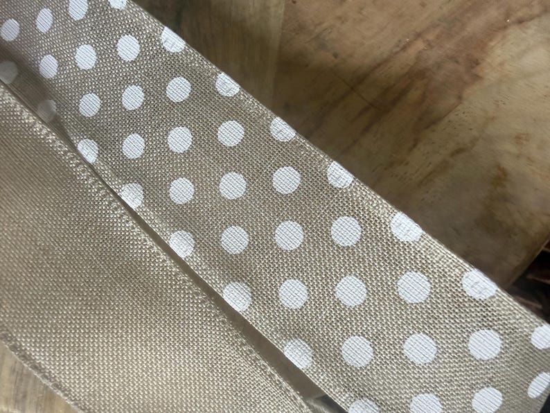 May include: Close-up of tan ribbon with white polka dots. The ribbon is made of a textured fabric and is folded over itself. The background is a wooden surface.