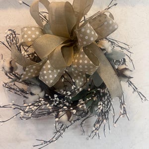 May include: A decorative wreath featuring cotton bolls, sprigs, and greenery. A large bow made of tan ribbon with white polka dots is centered on the wreath. The wreath is designed for home decor.