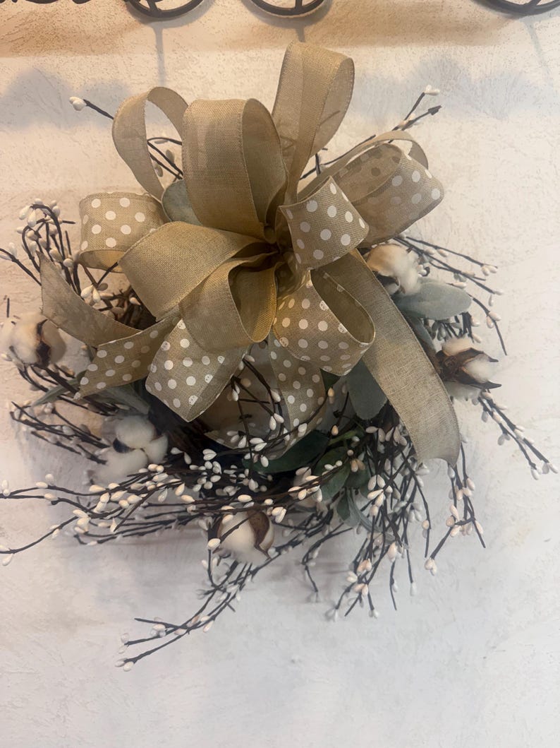 May include: A decorative wreath featuring cotton bolls, sprigs, and greenery. A large bow made of tan ribbon with white polka dots is centered on the wreath. The wreath is designed for home decor.