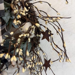 May include: A rustic wreath featuring a woven base adorned with faux cotton bolls, sprigs, and rusty metal star accents. The wreath includes a black and white buffalo check ribbon and muted green foliage, creating a farmhouse aesthetic.