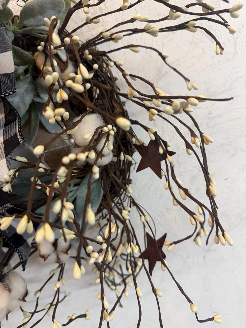 May include: A rustic wreath featuring a woven base adorned with faux cotton bolls, sprigs, and rusty metal star accents. The wreath includes a black and white buffalo check ribbon and muted green foliage, creating a farmhouse aesthetic.