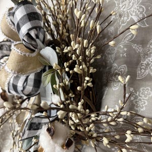 May include: A rustic wreath featuring a mix of natural elements. It includes a large bow with black and white checkered ribbon, burlap ribbon, and sprigs of white berries. A cotton boll adds texture.