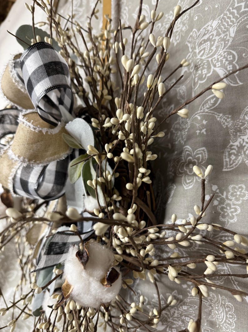 May include: A rustic wreath featuring a mix of natural elements. It includes a large bow with black and white checkered ribbon, burlap ribbon, and sprigs of white berries. A cotton boll adds texture.