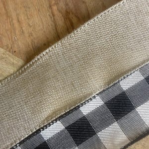 May include: Close-up of two ribbons. One is a natural tan burlap ribbon, and the other is a black and white checkered ribbon. The ribbons are angled across a wooden surface. The burlap ribbon has a woven texture, and the checkered ribbon has a smooth, glossy finish. Ideal for crafting.