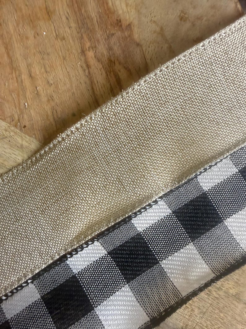 May include: Close-up of two ribbons. One is a natural tan burlap ribbon, and the other is a black and white checkered ribbon. The ribbons are angled across a wooden surface. The burlap ribbon has a woven texture, and the checkered ribbon has a smooth, glossy finish. Ideal for crafting.