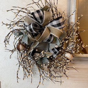 May include: A decorative wreath featuring a large bow with black and white checkered ribbon and natural burlap. The wreath is adorned with sprigs of white berries, cotton bolls, and green leaves, creating a rustic, farmhouse aesthetic.