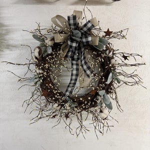 May include: A rustic grapevine wreath adorned with cotton bolls, sprigs, and faux greenery. A large bow with a black and white checkered pattern and a tan ribbon adds a decorative touch. Small metal stars are scattered throughout.