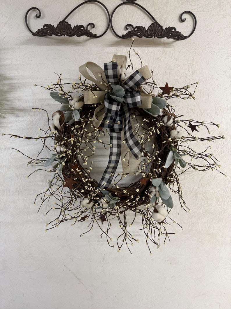 May include: A rustic grapevine wreath adorned with cotton bolls, sprigs, and faux greenery. A large bow with a black and white checkered pattern and a tan ribbon adds a decorative touch. Small metal stars are scattered throughout.