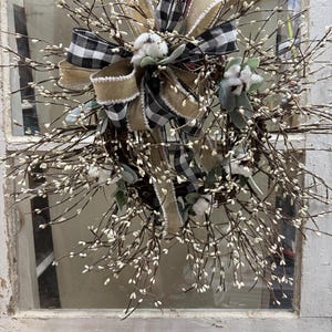 May include: A rustic wreath featuring a mix of natural elements. It has a base of brown twigs adorned with small white buds. A large bow made of black and white checkered fabric and tan burlap adds a decorative touch, along with cotton accents.