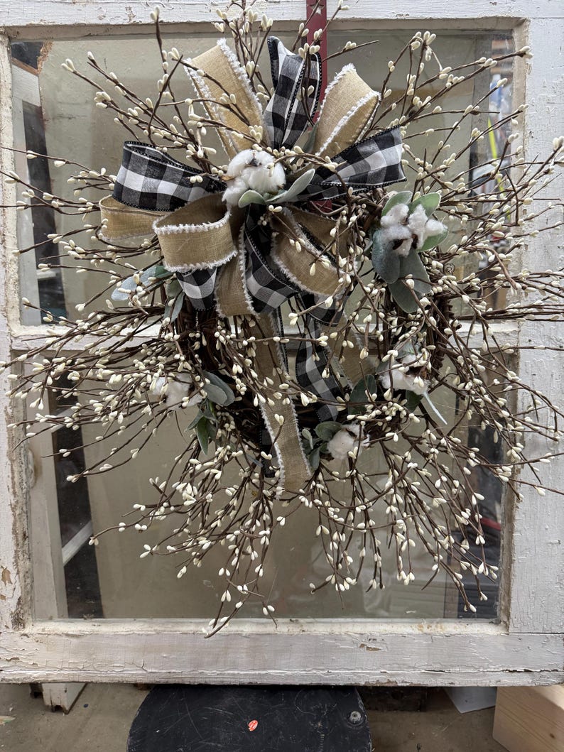 May include: A rustic wreath featuring a mix of natural elements. It has a base of brown twigs adorned with small white buds. A large bow made of black and white checkered fabric and tan burlap adds a decorative touch, along with cotton accents.