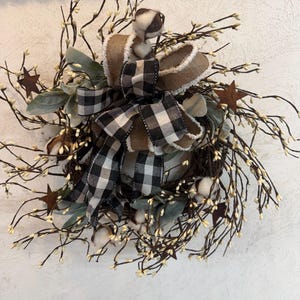 May include: A rustic wreath featuring a mix of natural elements. It includes brown twigs with small white buds, cotton bolls, and green leaves. A large bow with a black and white checkered pattern and a brown ribbon with a fluffy edge adds a decorative touch. Rusty star accents.