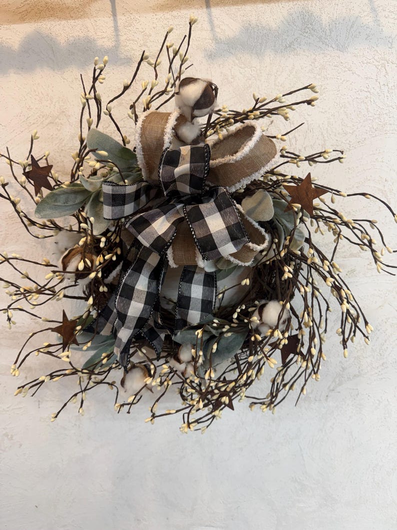 May include: A rustic wreath featuring a mix of natural elements. It includes brown twigs with small white buds, cotton bolls, and green leaves. A large bow with a black and white checkered pattern and a brown ribbon with a fluffy edge adds a decorative touch. Rusty star accents.