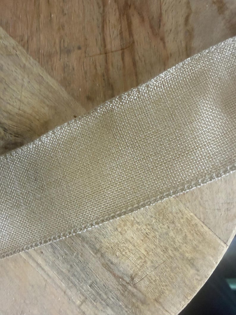 May include: A close-up of a gold mesh ribbon, likely for crafting or decoration. The ribbon has a fine, woven texture and a metallic sheen. The ribbon is positioned diagonally across a wooden surface.