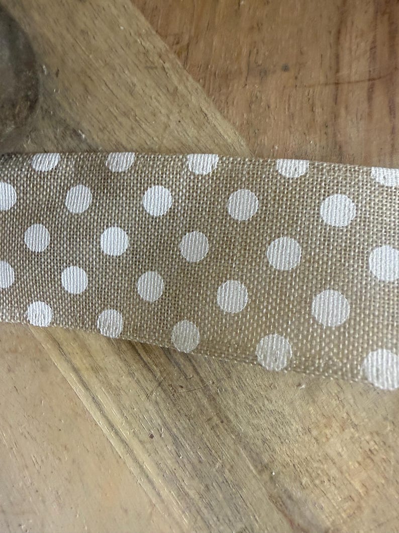 May include: Close-up of a tan burlap ribbon with a repeating pattern of white polka dots. The ribbon is lying on a wooden surface. The ribbon is likely used for crafts, gift wrapping, or decorative purposes.