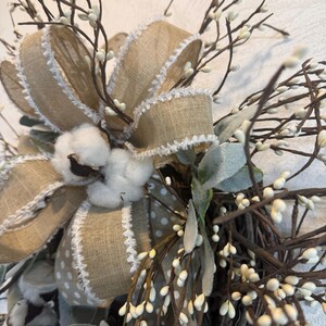 May include: Close-up of a decorative wreath featuring a large burlap bow with white trim and a polka dot pattern. The wreath includes cotton bolls, small white berries, and green leaves, creating a rustic, natural aesthetic.