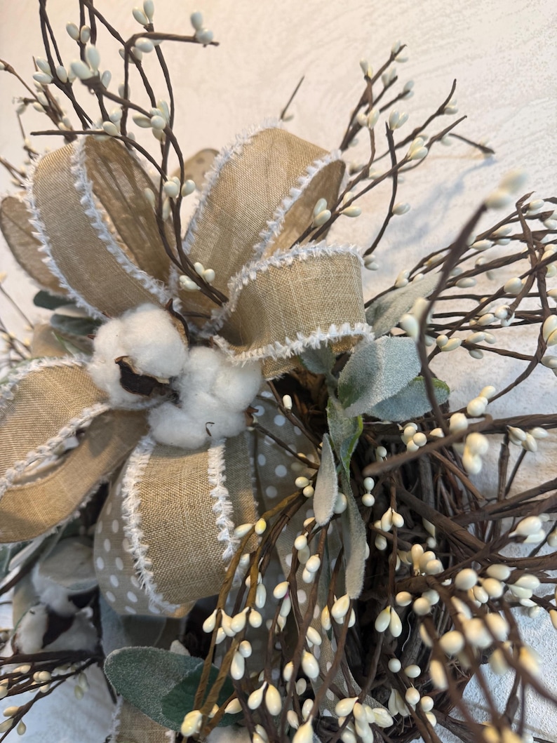 May include: Close-up of a decorative wreath featuring a large burlap bow with white trim and a polka dot pattern. The wreath includes cotton bolls, small white berries, and green leaves, creating a rustic, natural aesthetic.