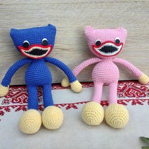 Puede incluir: Dos muñecos de ganchillo hechos a mano, uno azul y otro rosa, cada uno con una amplia sonrisa dentada. Los muñecos tienen pies y manos amarillos y están colocados sobre una superficie estampada. Los muñecos son un diseño juguetón y caprichoso.