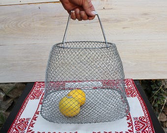 May include: A vintage wire mesh basket with a metal handle, holding two yellow lemons. The basket is set on a table with a red and white patterned cloth. The background features a wooden panel and stone wall.
