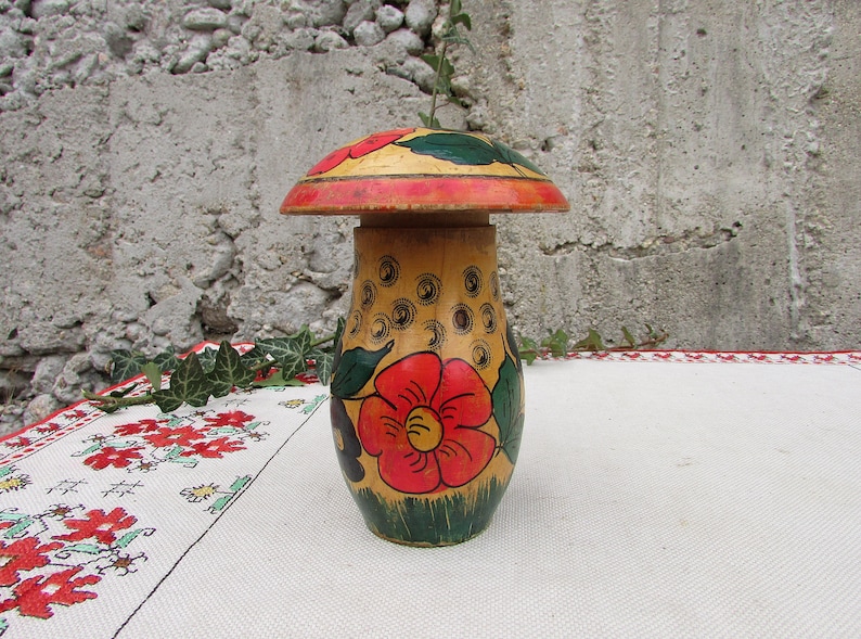 May include: A hand-painted wooden mushroom-shaped container with a red, yellow, and green floral design. The container is closed with a lid that is also painted with a floral design.