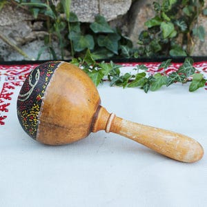 Pode incluir: Uma maraca de madeira com uma cabeça decorada em forma de cabaça. A cabeça é pintada com um padrão escuro e pontos amarelos e vermelhos. O cabo é de cor castanho claro. A maraca repousa sobre um tecido branco com uma borda bordada a vermelho.