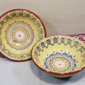 Puede incluir: Dos cuencos de cerámica con fondo amarillo y complejos diseños florales en naranja, azul y verde. Los cuencos tienen un borde marrón y están colocados sobre una superficie blanca con un textil estampado en rojo y blanco.