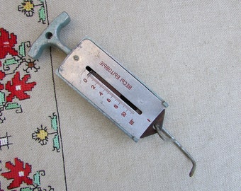 May include: A silver spring scale with a black handle and a hook. The scale has a dial with red markings and the text "BECI БРИТОВБIE" printed on it.