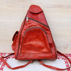 Puede incluir: Una mochila de cuero roja con una sola correa. La mochila tiene una solapa con cierre de cremallera y un bolsillo grande en la parte delantera.