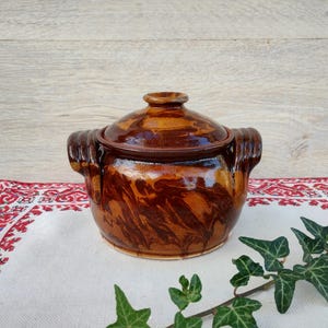 Op de afbeelding: Een bruine keramische pot met een deksel, met een marmerpatroon. De pot heeft twee handvatten en staat op een wit tafelkleed met rode borduurwerk.