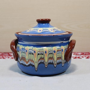 May include: A blue ceramic pot with a lid and two brown handles. The pot features a decorative pattern in yellow, orange, and white. The lid has a brown knob. The pot is sitting on a white surface with a red and white patterned cloth.