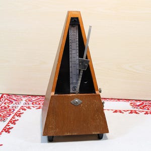 Può includere: Un metronomo in legno a forma di piramide. Il metronomo ha una finitura in legno scuro e un pendolo argentato. La faccia del metronomo ha una scala con numeri. Il metronomo è su una superficie bianca con un panno a motivi rossi e bianchi.