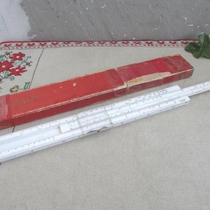 May include: A vintage white slide rule with black markings and a red rectangular case. The slide rule is open, displaying its scales and indicators. The case is made of cardboard and shows signs of wear.