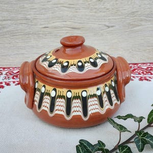 Puede incluir: Una olla de cerámica marrón con tapa, con un diseño decorativo de patrones negros, blancos y amarillos. La olla tiene dos asas y un pomo en la tapa. Está sobre una superficie blanca con un paño estampado en rojo y blanco.