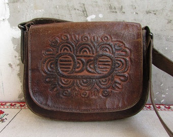 May include: A brown leather shoulder bag with an intricate embossed design. The bag has a flap closure and a long adjustable strap.