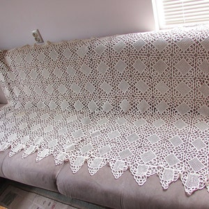 Pode incluir: Uma toalha de mesa de crochê branca com um padrão de diamantes e bordas festoneadas. A toalha de mesa está drapeada sobre um sofá marrom.