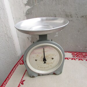 Op de afbeelding: Een vintage grijze en witte keukenweegschaal met een zilveren weegschaal. De weegschaal heeft een ronde wijzerplaat met zwarte cijfers en een zwarte wijzer. De wijzerplaat heeft ook de tekst "Echt" en "Campina" in zwarte letters. De weegschaal staat op een wit tafelkleed met een rode geborduurde rand.