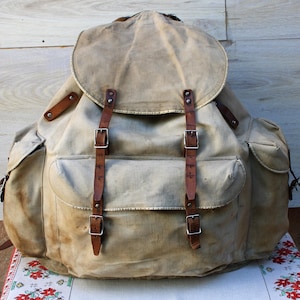 Può includere: Uno zaino in tela beige dotato di una grande patta superiore, due tasche laterali e una tasca frontale centrale. La borsa è fissata con due cinghie verticali in pelle marrone dotate di fibbie e rivetti in metallo argentato per la chiusura.