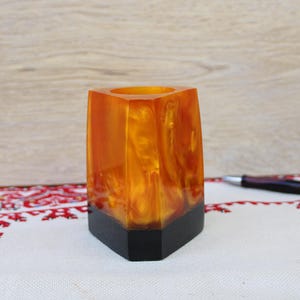 May include: A geometric pen holder with a black base and a translucent amber-coloured upper section. The pen holder is made of resin and has a unique, swirled pattern. A pen rests on the patterned surface.