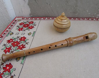 Puede incluir: Un flautín de madera con seis agujeros para los dedos y una caja de madera con tapa y tallas intrincadas. El flautín y la caja están sobre un mantel blanco con un patrón floral rojo y verde.
