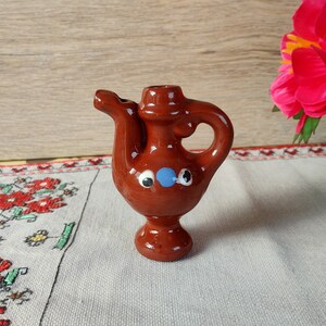 Può includere: Un piccolo teiera in ceramica marrone con manico e beccuccio. Ha un viso dipinto di blu e bianco con due occhi e un naso.