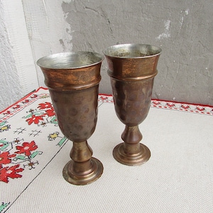 May include: Two antique copper goblets with hammered metal surfaces. The goblets have a rounded bowl and a flared base.