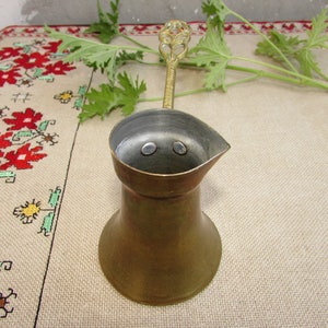 Peut inclure: Une cafetière turque en cuivre avec une poignée décorative. La cafetière est posée sur une nappe blanche à motif floral rouge et vert.