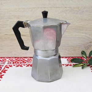 May include: A silver stovetop espresso maker with a black handle and lid knob. The coffee pot sits on a white and red patterned cloth, with a sprig of green leaves to the side. The background is a light wood.