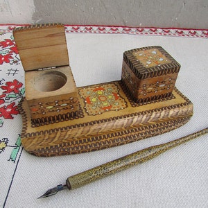 May include: A wooden inkwell set with a small inkwell, a larger inkwell with a lid, and a wooden pen. The set is decorated with intricate floral patterns and a geometric design.
